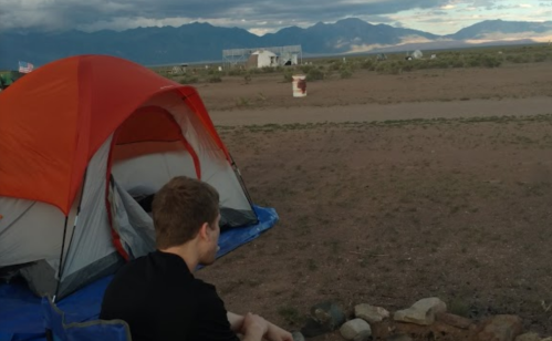 A person sits near a tent in a desert landscape, with mountains in the background and a cloudy sky.