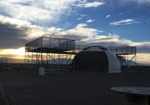 A dome structure on a construction site with metal scaffolding, set against a sunset and cloudy sky.
