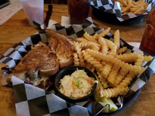 A plate with a grilled sandwich, crinkle-cut fries, coleslaw, and a pickle, served in a checkered basket.