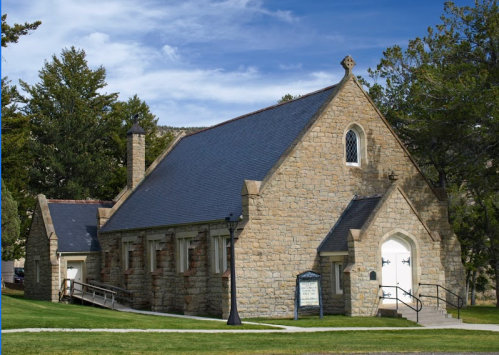 A stone church with a steep roof, surrounded by trees and a clear blue sky.