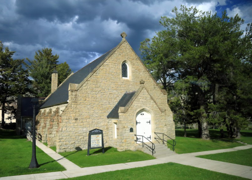 A small stone church with a white door, surrounded by green grass and trees, under a cloudy sky.