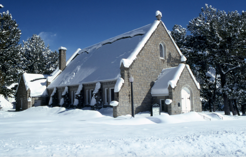 A stone building covered in snow, surrounded by trees, under a clear blue sky.