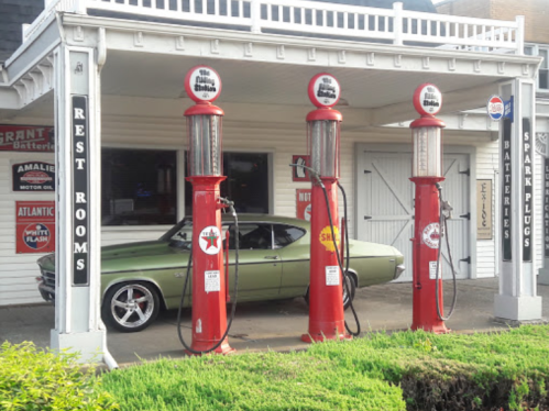 A vintage gas station with three red pumps and a classic green car parked in front.