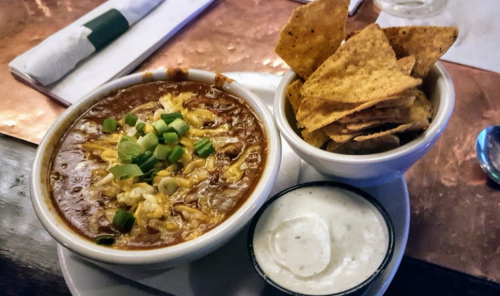 A bowl of chili topped with cheese and green onions, served with a side of tortilla chips and a small bowl of dip.