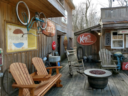 A rustic outdoor space with wooden chairs, a fire pit, and vintage soda signs, creating a cozy, nostalgic atmosphere.