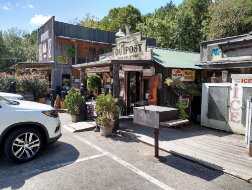 A rustic general store with wooden signage and a porch, surrounded by greenery and parked cars.