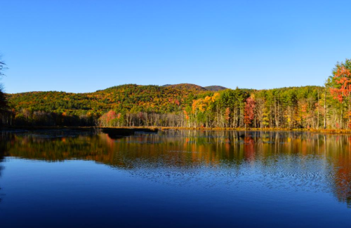 A serene lake reflecting colorful autumn trees and a clear blue sky, surrounded by rolling hills.