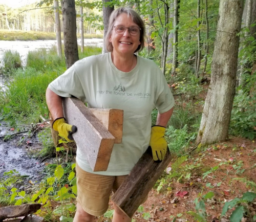 A smiling woman in a light green shirt and gloves carries two pieces of wood in a forested area by a pond.