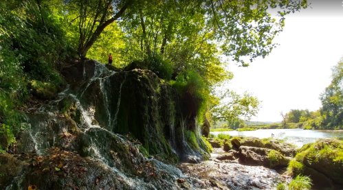 A serene waterfall cascades over rocks, surrounded by lush greenery and a calm river under a bright sky.