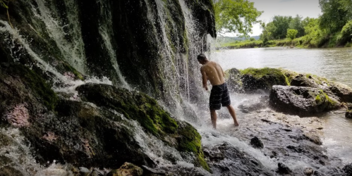A person stands under a waterfall, surrounded by rocks and greenery, with water cascading down around them.