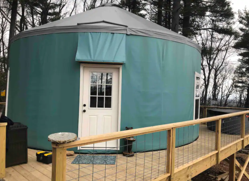 A teal yurt with a white door, situated on a wooden deck surrounded by trees.