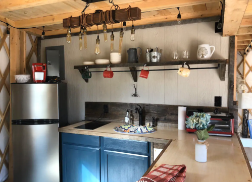 Cozy kitchen with stainless steel fridge, blue cabinets, wooden shelves, and decorative lighting.