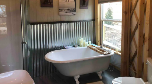 A cozy bathroom featuring a freestanding tub, corrugated metal wall, candles, and natural light from a window.