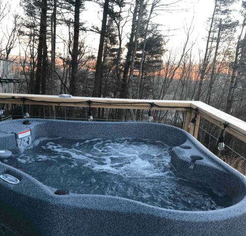A hot tub on a wooden deck surrounded by trees, with a sunset visible in the background.