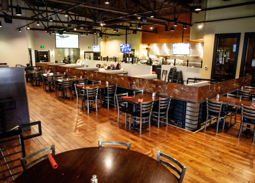 Interior of a modern restaurant with wooden floors, tables, and a bar area, featuring a casual dining atmosphere.