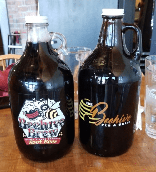Two glass growlers of root beer, one labeled "Beehive Brew" and the other "The Beehive Pub & Grill."