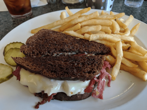 A plate with a sandwich on rye bread, filled with meat and mayo, accompanied by fries and pickles.