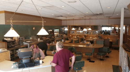 A cozy diner interior with green booths, a counter, and patrons enjoying their meals. Warm lighting creates a welcoming atmosphere.