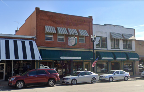 Historic brick storefronts with awnings, parked cars, and an American flag in a small town setting.