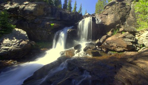 A serene waterfall cascades over rocky terrain, surrounded by lush greenery and clear blue skies.