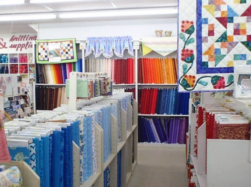 A fabric store aisle filled with colorful rolls of fabric and quilting supplies, with quilts displayed on the walls.