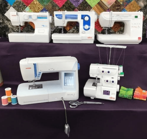 A display of sewing machines and a serger, with colorful spools of thread and scissors on a purple tablecloth.