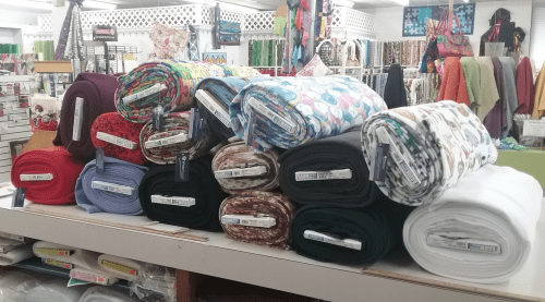 A display of neatly rolled fabric bolts in various colors and patterns stacked on a table in a fabric store.
