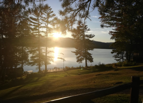 Sunset over a calm lake, framed by trees, with a reflection shimmering on the water's surface.