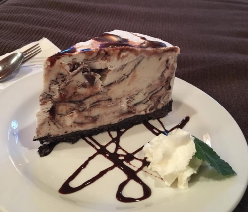 A slice of chocolate marble cheesecake on a plate, drizzled with chocolate sauce and served with whipped cream and mint.