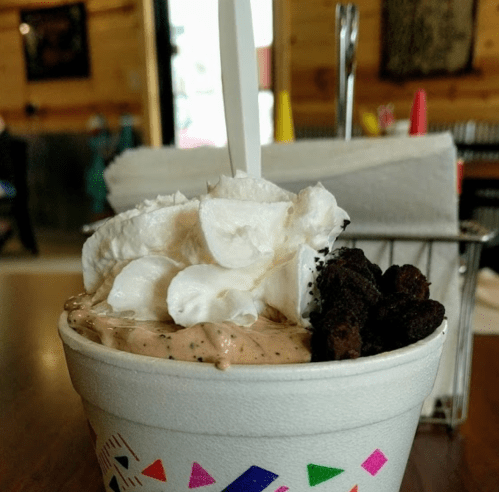 A cup of ice cream topped with whipped cream and cookie crumbles, set on a wooden table in a casual dining space.