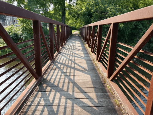 A wooden bridge with metal railings spans a river, casting shadows on the path under bright sunlight.