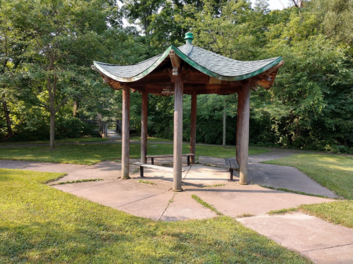 A wooden gazebo with a green roof, surrounded by grass and trees in a park setting.