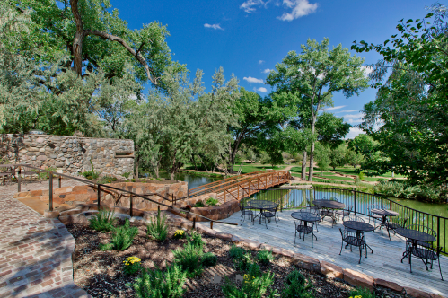 A scenic outdoor area with a wooden bridge, stone walls, and tables by a pond, surrounded by lush greenery and blue skies.