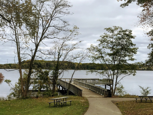 A serene lake view with a wooden pier, surrounded by trees and a grassy area with picnic tables.