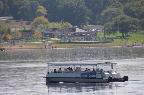 A boat named "Island Queen" sails on a lake, with a beach and park visible in the background.