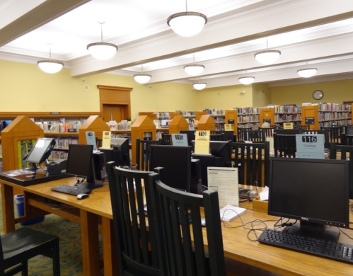 A quiet library interior with rows of computers and bookshelves, featuring wooden tables and overhead lighting.
