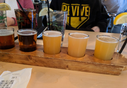 A wooden flight tray with five small glasses of different craft beers, alongside two larger drinks in the background.