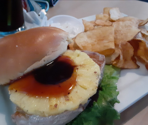 A burger with a pineapple slice and sauce, served with a side of potato chips and a small cup of dipping sauce.