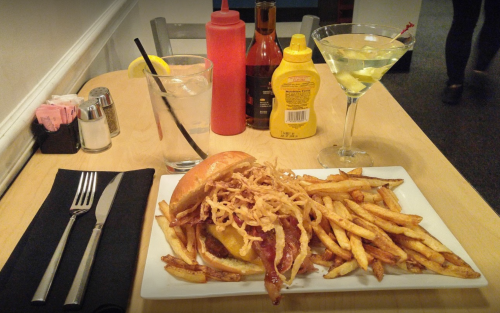 A plate with a burger topped with bacon and fried onions, served with fries, alongside drinks and condiments on a table.