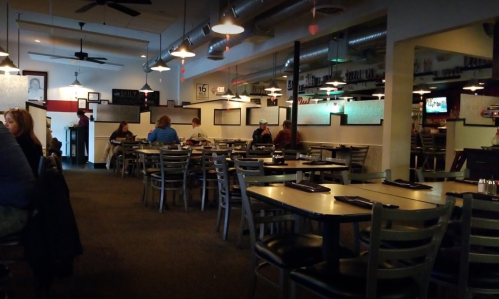 A spacious restaurant interior with tables, chairs, and patrons dining, featuring a bar area in the background.