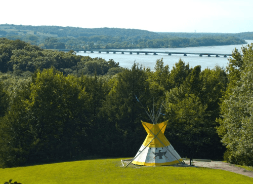 A colorful teepee stands on green grass, overlooking a river and a distant bridge surrounded by trees.