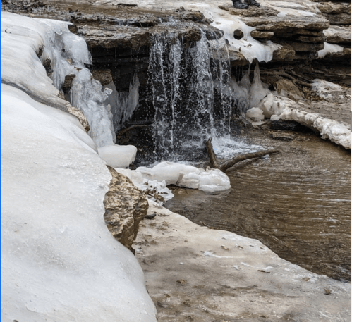 A small waterfall flows over icy rocks, surrounded by snow and frozen ground.
