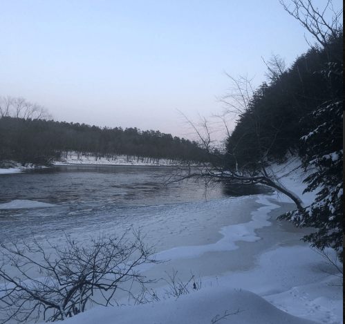 A serene winter landscape featuring a frozen river, snow-covered banks, and bare trees against a pastel sky.