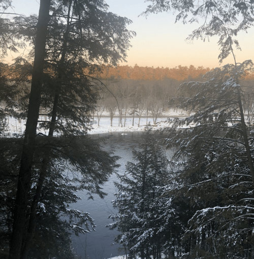 A serene winter landscape featuring a river surrounded by snow-covered trees and a soft sunrise glow in the background.