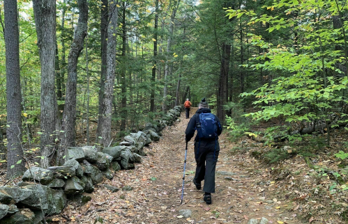 Two hikers walking along a forest trail lined with stone walls, surrounded by trees and autumn foliage.