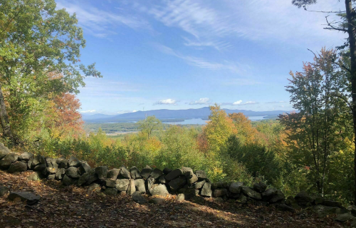 Scenic view of a colorful autumn landscape with a stone wall, mountains, and a lake under a blue sky.