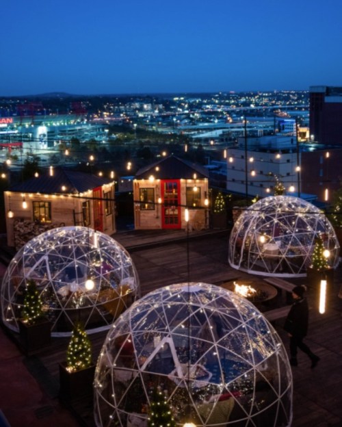 Rooftop scene with illuminated igloos, string lights, and a city skyline at dusk. Cozy winter atmosphere.