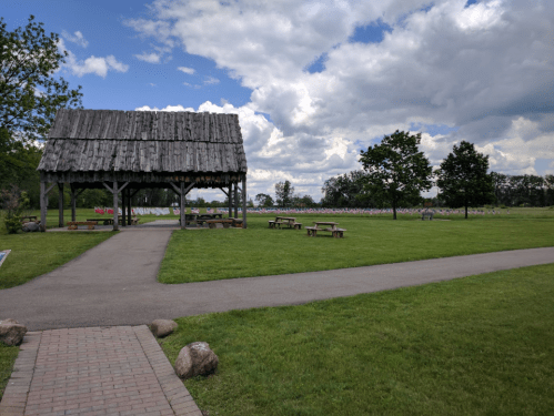 A grassy area with a wooden pavilion, picnic tables, and a path, under a partly cloudy sky.