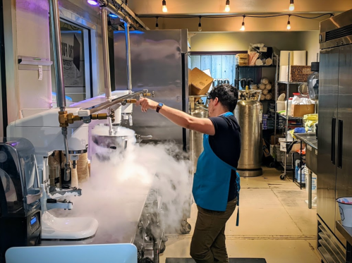 A person in an apron uses a machine, creating a cloud of vapor in a busy kitchen filled with equipment and supplies.