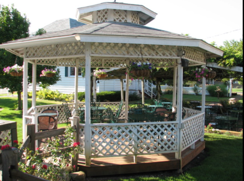 A white gazebo with lattice walls, surrounded by green grass and hanging flower baskets, offers a cozy outdoor seating area.